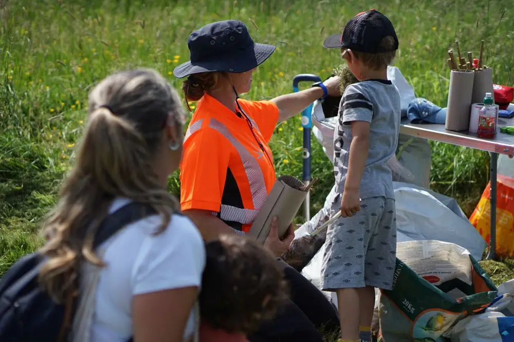 A woman in an orange safety vest helps a child at an outdoor table, whilst another woman and small child stand nearby in a grassy field. Gardening supplies are on the table. | Microsite | Microsite